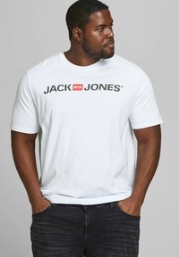 Weißes Baumwoll-T-Shirt mit einem auffälligen schwarzen "JACK & JONES" Logo in einem roten Kästchen, Rundhalsausschnitt und kurzen Ärmeln, kombiniert mit dunklen Jeans.