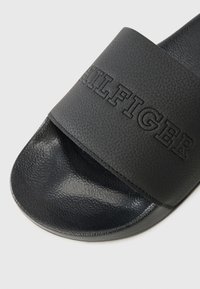 Black slide sandal with a textured faux leather strap featuring embossed brand name. Smooth sole with rounded edges for comfort.