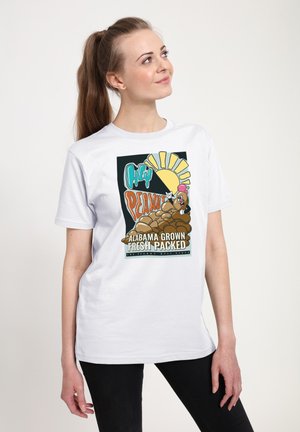 Kvinde med lys hud og brunt hår i en hestehale, der bærer en hvid t-shirt med en farverig jordnødde-tegneserie og teksten "Alabama grown fresh packed".