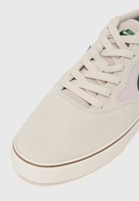 Sneaker din suede bej cu o textură netedă, șireturi plate și un logo verde distinctiv. Talpă din cauciuc albă cu un accent maroniu pe margine.