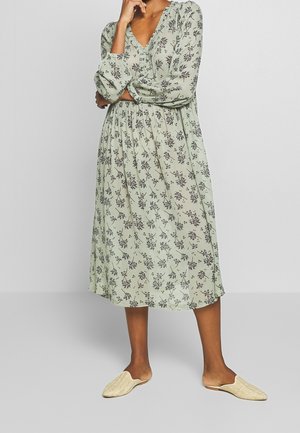 Femme portant une robe midi fleurie à manches longues vert clair et des chaussures tissées beige à enfiler, debout avec un bras croisé et la main près du visage.