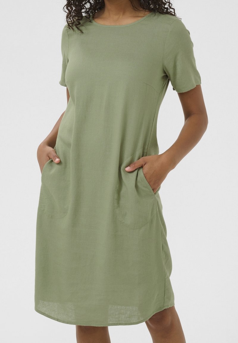 Vrouw in een licht olijfgroen, knielang jurkje met korte mouwen en handen in de voorzakken, tegen een effen achtergrond.