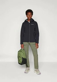 Czarna wodoodporna kurtka z zamkiem, zielone spodnie i zielony plecak z czarnymi akcentami. Szare sneakersy dopełniają stój.