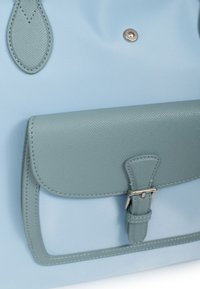 Borsa azzurra chiara con dettagli in pelle di un verde chiaro. Presenta un bottone a scatto e una patta testurizzata con dettaglio della fibbia. Superficie in tessuto liscia visibile.