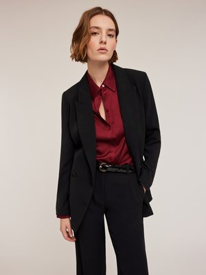 Zwarte blazer over een bordeaux satijnen shirt met een knopsluiting. Textuurstof en een glanzende zwarte riem maken de look compleet.