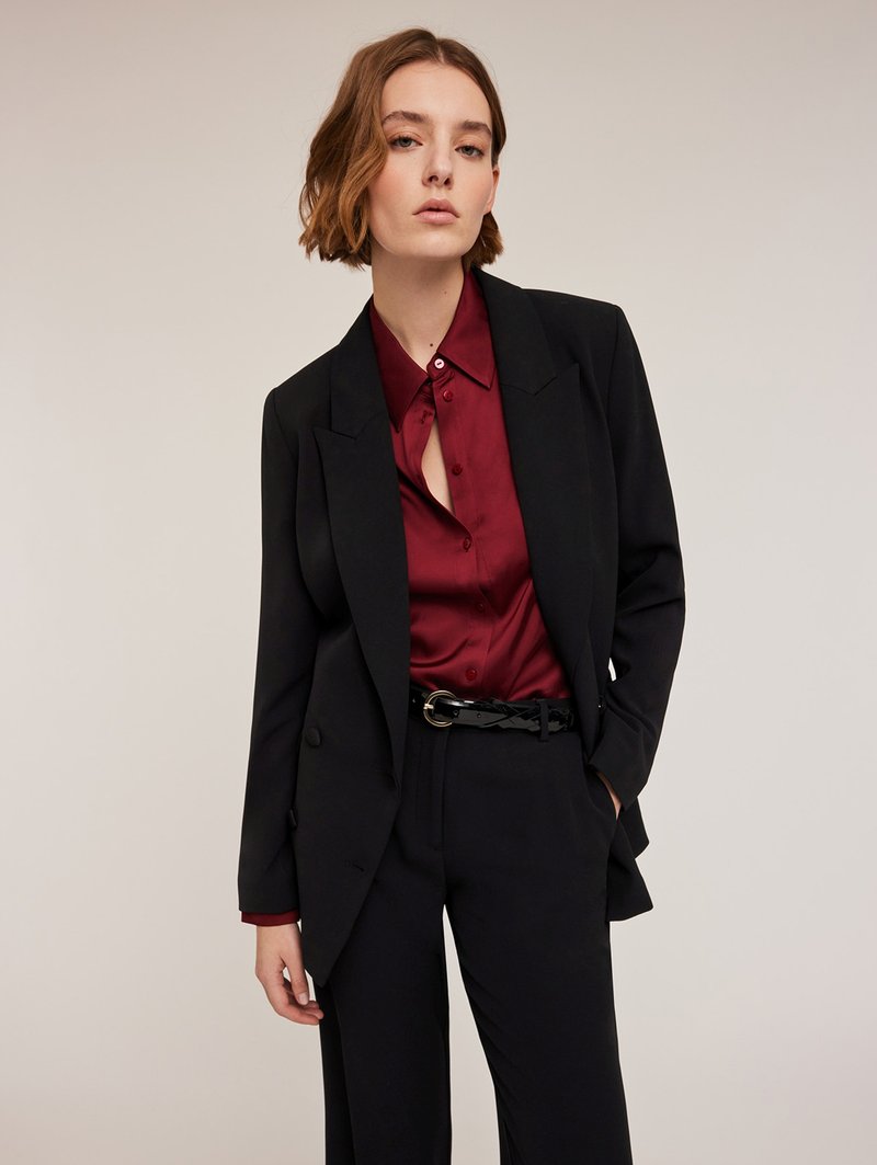 Zwarte blazer over een bordeaux satijnen shirt met een knopsluiting. Textuurstof en een glanzende zwarte riem maken de look compleet.