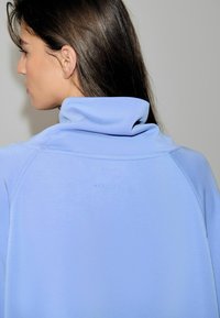 Lyseblå sweatshirt med høj krave, blød tekstur og diskret mærkning på bagsiden. Har raglanærmer for nem bevægelse.