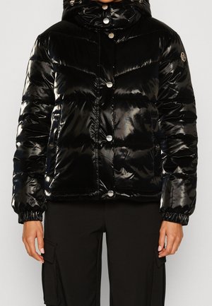 Giacca a vento imbottita nera lucida con cappuccio, bottoni a pressione frontali, tasche laterali, polsini elasticizzati, indossata con pantaloni cargo neri.