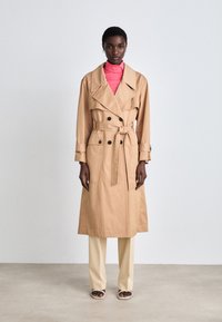Beige trenchcoat met brede revers, dubbele knopen en een strikceintuur. Gecombineerd met een roze top en crèmekleurige broek. Zachte textuur.