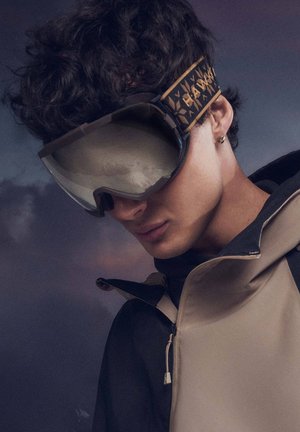 Gafas de esquí con una lente grande y curva, montura oscura y correa con estampado. El modelo lleva una chaqueta bicolor; la capucha está levantada, dejando al descubierto la barbilla.