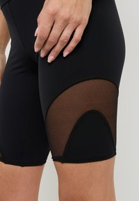 Pantaloni corti neri traspiranti con pannelli in mesh trasparente sui lati e design aderente, con dettagli di cucitura nera a contrasto.