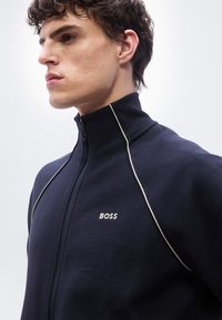 Νεαρός άνδρας με κοντά σγουρά μαλλιά που φοράει navy μπλε ζακέτα με φερμουάρ και λευκές ρίγες, με το λογότυπο "BOSS" στο στήθος, στραμμένος προς τα αριστερά.