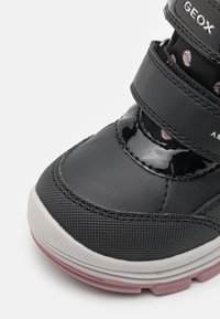 Černé tenisky se syntetickým a strukturovaným materiálem, s suchým zipem, lesklými detaily a gumovou podrážkou s nádechem růžové.