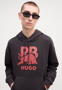 Svart hettegenser med en rød "RB" logo og okse-grafikk, med snorhett og kengurulomme. Modellen har på seg mørke solbriller.