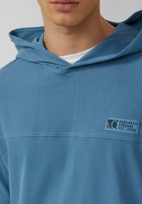 Jasnoniebieska bluza z kapturem wykonana z bawełnianej mieszanki tkanin, z przednią kieszenią, regulowanym kapturem ze ściągaczem oraz małą naszywką z logo i napisem.