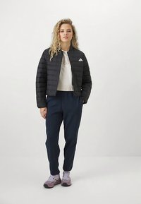 Γυναίκα με σγουρά μαλλιά που στέκεται, φορώντας ένα μαύρο μπουφάν, λευκό κοντό μπλουζάκι, ναυτικές φόρμες και ανοιχτό μωβ αθλητικά παπούτσια μπροστά από ένα λευκό υπόβαθρο.