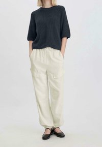 Femme portant un pull en maille noir à manches mi-longues, un pantalon cargo crème avec taille à cordon, et des chaussures Mary Jane noires.