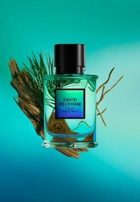 Transparent glasparfymflaska med svart kork, med en iriserande etikett som läser "David Beckham True Instinct" mot en teal bakgrund.