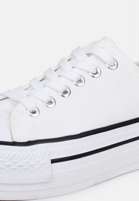 Sneaker bianca in tela con un design low-profile, caratterizzata da occhielli metallici, lacci bianchi e un accento nero sulla spessa suola in gomma.