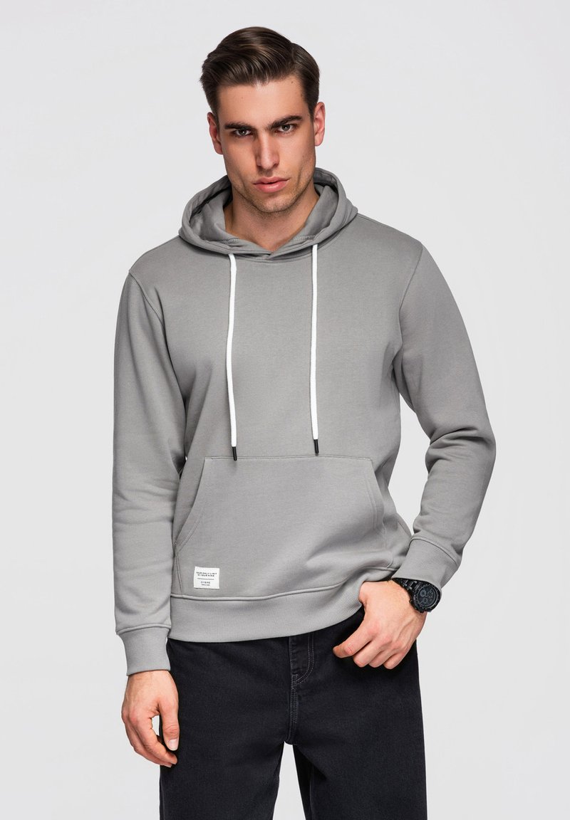 Jeune homme aux cheveux courts portant un sweat à capuche gris avec des cordons blancs et un pantalon noir, posant avec une main près de la poche.