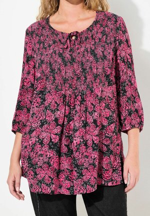 Kobieta w luźnej czarnej bluzce z jasnoróżowym kwiatowym wzorem, rękawami o długości do łokcia oraz małym wiązaniem z przodu przy dekolcie.