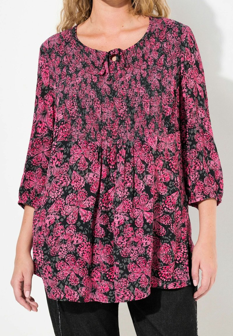 Femme portant un chemisier noir ample avec un motif floral rose vif, manches trois-quarts, et un petit lien à l'avant au niveau du col.