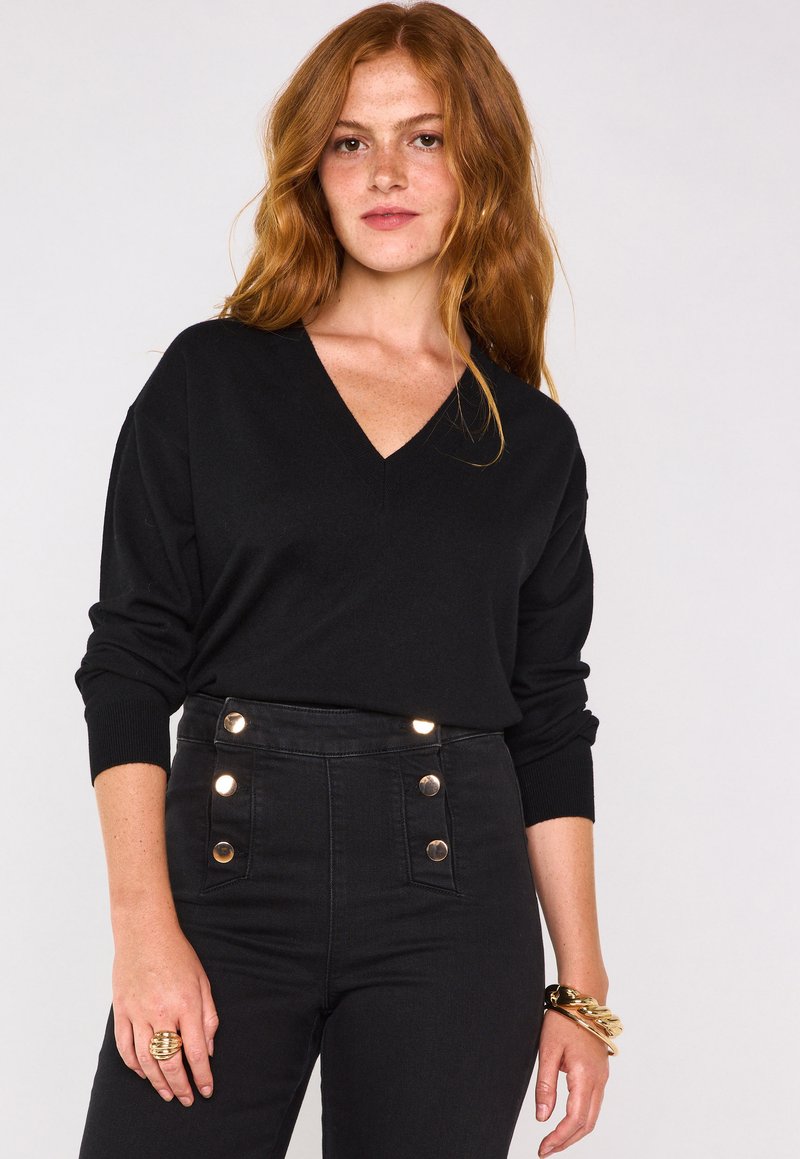 Maglione nero a V con maniche lunghe, abbinato a jeans neri a vita alta con bottoni dorati e un anello testurizzato alla mano.