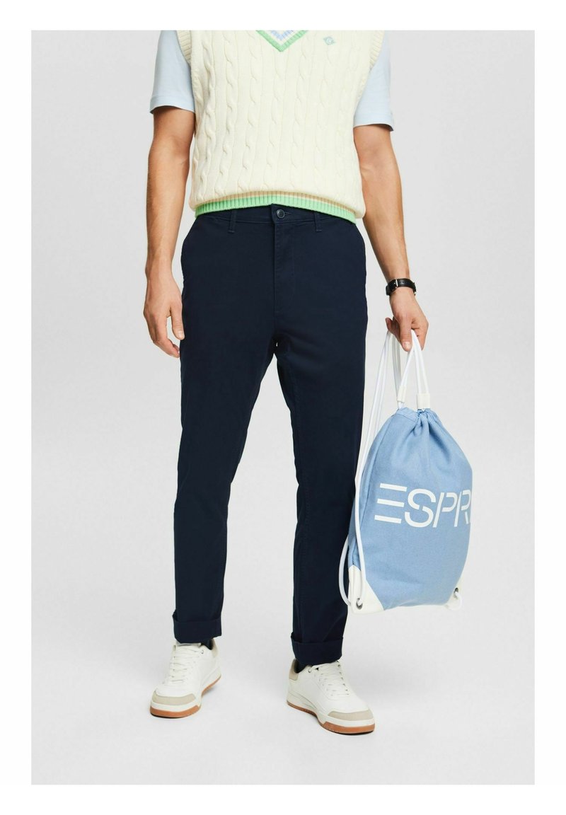 Homme portant un pantalon bleu marine, des baskets blanches, un gilet en maille torsadée crème par-dessus une chemise bleu clair, tenant un sac à cordon ESPRIT bleu clair.