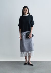 Crni oversized pleteni džemper s kratkim rukavima, upareni s sivom suknjom duljine do koljena s bijelom čipkanom obrubom. Drži crnu teksturiranu torbicu.