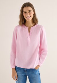 Femme souriante portant un chemisier rose clair à manches longues avec une encolure fendue et un jean bleu, debout devant un fond beige.