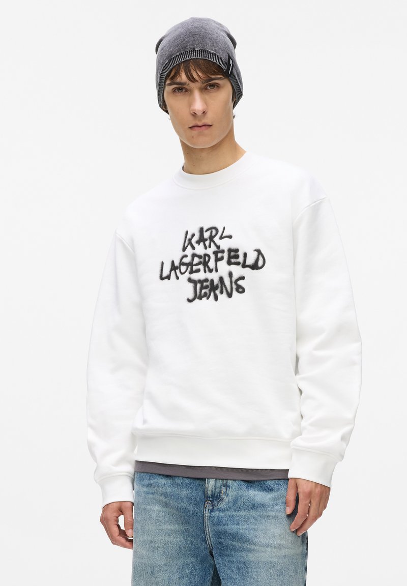 Bijela majica s kapuljačom s crnim grafitnim tekstom "KARL LAGERFELD JEANS" na prednjoj strani. Uparena sa izblijedjelim plavim trapericama i sivim šeširom.