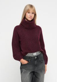 Pullover bordeaux con collo a dolcevita e maniche ampie, di texture lavorata a maglia, abbinato a jeans neri sbiaditi a vita alta con una cintura a quadri.
