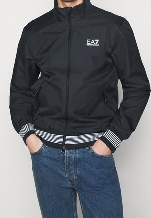 Osoba ubrana w czarną kurtkę na zamek EA7 Emporio Armani z paskami na mankietach i dole, ręce w kieszeniach, w połączeniu z niebieskimi dżinsami.