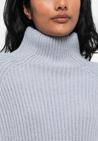 Maglione a collo alto in maglia a coste di un grigio chiaro, dal taglio rilassato, che mostra una texture morbida e dettagli di cucitura sottili.