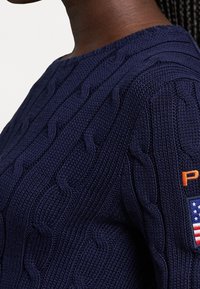 Maglione blu navy lavorato a maglia con motivo a trecce testurizzato, scollo tondo e dettaglio logo arancione con toppa della bandiera americana sulla manica.