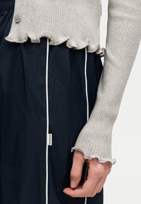 Κοντινό πλάνο ενός χεριού που φοράει ένα γκρι ανοιχτό πλεκτό πουλόβερ με ριγωτές ραφές και βολάν στα μανίκια πάνω από ένα μαύρο ένδυμα με λευκές κάθετες γραμμές και μια μικρή ετικέτα.