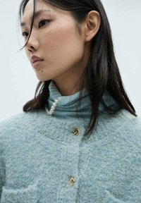 Maglione di lana azzurro chiaro con texture e colletto alto, impreziosito da bottoni dorati e una collana di perle decorativa. Aspetto morbido e accogliente.