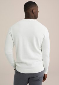 Witte gebreide trui met een gestructureerde weef, ronde halslijn en ribgebreide boorden en zoom, gedragen met grijze broek, van achteren gezien.