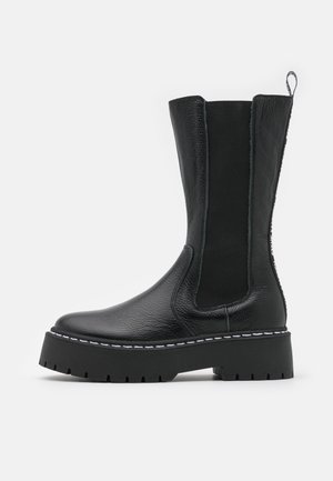 Schwarzer knöchelhoher Lederstiefel mit grober Profilsohle, elastischen Seitenteilen, weißen Nähten und einer Zuglasche hinten.
