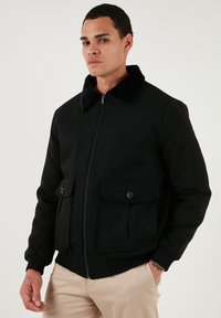 Sort bomberjakke lavet af uldblanding, med en falsk pelskrave, to frontlommer og ribbede manchetter og kant. Lynlås lukning.