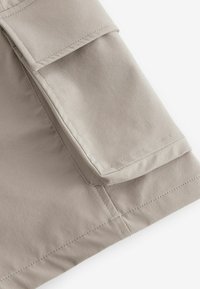 Un tissu beige avec une texture lisse présente une grande poche à rabat. La couture est soigneusement réalisée, mettant en valeur le design épuré.