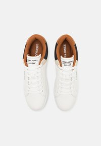 White sneakers made of smooth material with tan and black accents, featuring round laces and branding on the tongue and side.