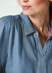 Hellblaues Denimhemd mit Kragen, gefalteten Schultern und weicher Textur. Die vordere Knopfleiste ist teilweise geöffnet, wodurch der Glanz des Stoffes sichtbar wird.