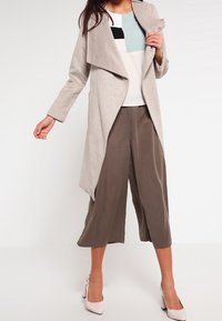 Cappotto lungo grigio chiaro con apertura frontale, indossato sopra un top a blocchi di colore. Pantaloni ampi marrone scuro e scarpe con tacco alto rosa chiaro.