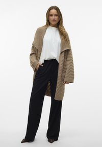 Beige Strickcardigan mit gerippter Textur, kombiniert mit einem weißen T-Shirt und schwarzen weiten Hosen. Braune Schuhe vervollständigen das Outfit.