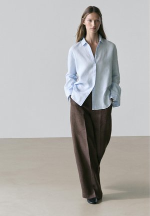 Frau, die einen hellblauen, übergroßen Hemd mit hochgekrempelten Ärmeln und weiten braunen Hosen trägt, läuft vor einfachem neutralem Hintergrund.