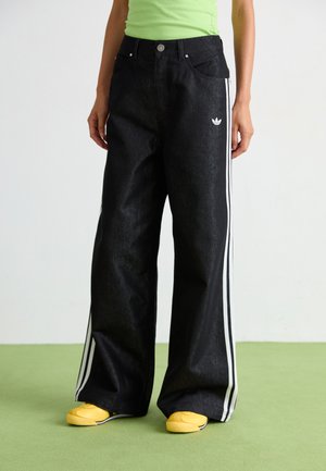 Pantalons large en denim noir avec des bandes blanches sur les côtés et un logo brodé à l'avant, associés à des baskets jaunes. Texture lisse.