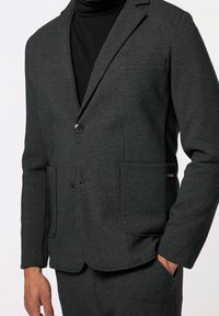 Muž v tmavě šedém saku s dvěma předními kapsami a černém roláku, s jednou rukou v kapse kalhot.