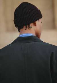Sort ribbet strikket beanie, blå skjorte med krave lagd under en mørk jakke, der fremhæver rene linjer og et enkelt design. Teksturerede stofdetaljer synlige.