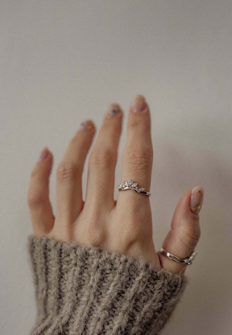 Main avec deux bagues en argent sur le majeur et le pouce, portant un pull en maille beige, avec un vernis à ongles de couleur neutre et une légère décoration des ongles.
